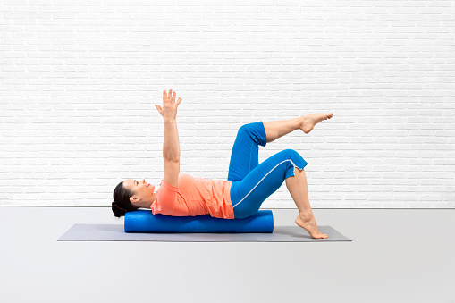 Fitness recovery. Adult woman lying on back along foam roller and practice classic pilates exercise in studio, indoor. Abs and core drill, balance, recreation, workout, wellness concept