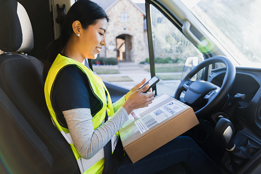 Still sitting in the driver's seat, the young adult female delivery person uses a digital tablet to scan the barcode on a box she is about to drop off at an address in a residential neighborhood.