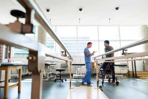 A female physical therapist takes notes before assisting a young man with walking after a leg injury. Parallel bars are in the foreground.