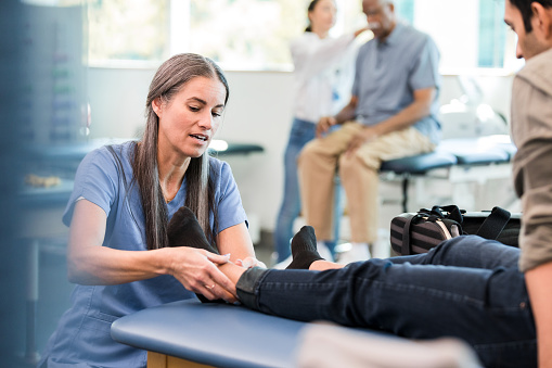 A female physical therapist examines a male patient's ankle during a physical therapy session.