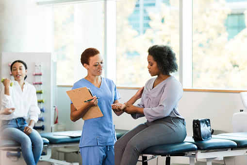 While a young adult woman exercises with hand weights in the background, the female mid adult occupational therapist asks the mid adult businesswoman about the pain in her wrist.