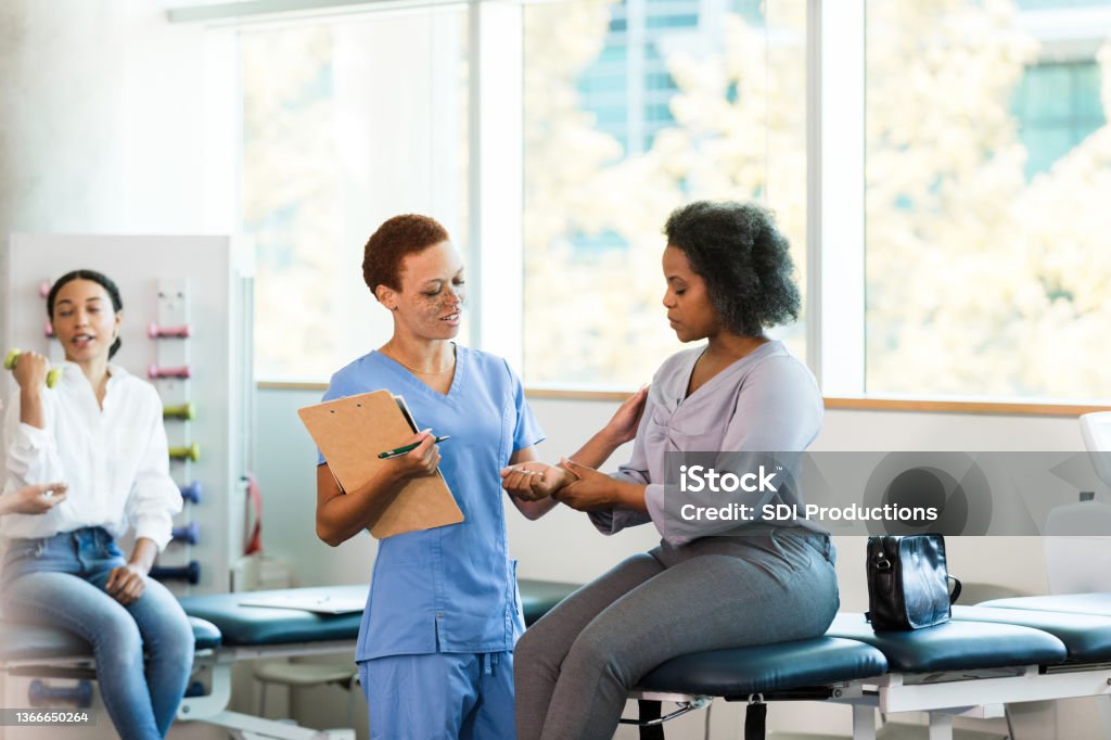 Female occupational therapist asks businesswoman about wrist pain While a young adult woman exercises with hand weights in the background, the female mid adult occupational therapist asks the mid adult businesswoman about the pain in her wrist. Healthcare And Medicine Stock Photo