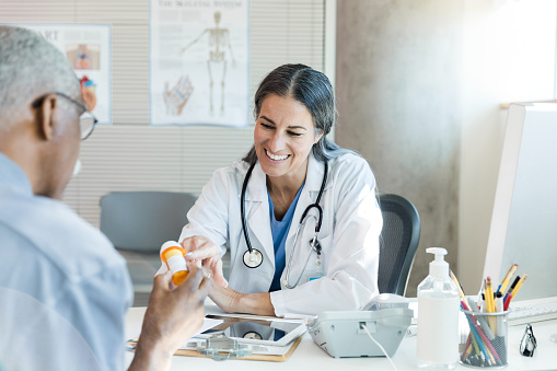 The female mid adult doctor smiles as she reaches across the desk to explain medication side effects to the unrecognizable male senior adult patient.  The doctor points to the pill bottle held by the patient.