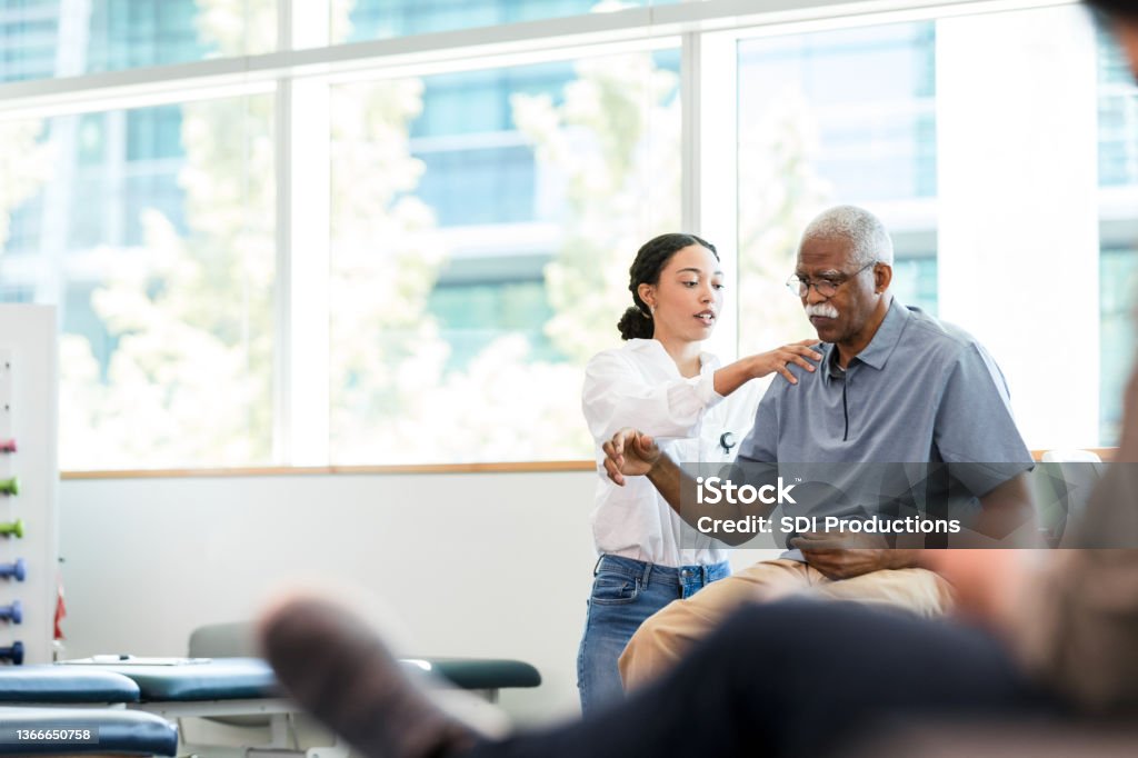 Physical therapist examines senior man's shoulder As the senior man grimaces, the young adult female physical therapist manipulates his injured shoulder. Physical Therapy Stock Photo