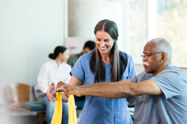Therapist smiles as senior man learns to use elastic band As other therapists work with patients in the background, the mid adult female physical therapist teaches the senior adult man how to use an elastic band to exercise his arms. occupational therapy photos stock pictures, royalty-free photos & images