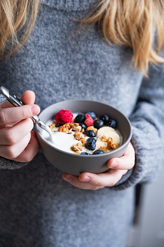 a young girl in a gray sweater is holding a bowl of vegetarian yogurt, granola and fresh blueberry and banana. The concept of a healthy breakfast.