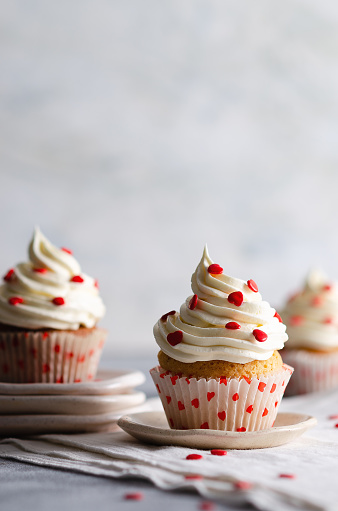 Vanilla muffins with buttercream frosting sprinkled with red sugar hearts on plates and a white cloth, on a grey backdrop.