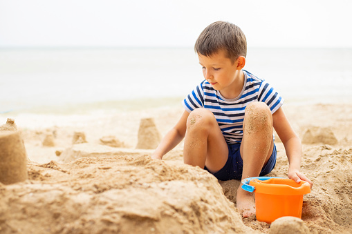 Happy child playing with sand on the beach in summer. Boy builds a sand castle on the sea.