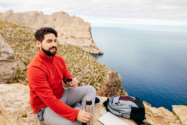 Young hiker sitting with a laptop and his backpack enjoying a hot tea from a thermos bottle with a smile on a rocky wall with the sea of the bay of Pollensa on the north coast of Mallorca in the background. Intended for life-balance and workation concepts. Color editing. Part of a series.