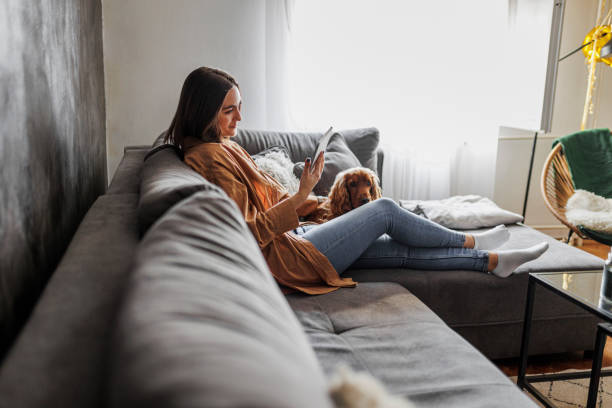 mujer deprimida que usa la tableta digital y descansa con su perro en casa - mujer leyendo periodico fotografías e imágenes de stock