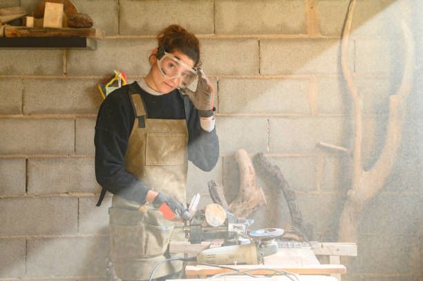 Craftswoman wearing apron and gloves takes off protective goggles while working in a wood workshop. Portrait of young professional woman wearing goggles working in a carpentry workshop against wood. Confident engineer. carpenter carpentry craftsperson carving stock pictures, royalty-free photos & images