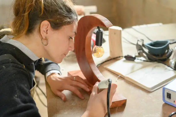 Photo of Beautiful young craftswoman engraving on handmade wooden lamp while using professional instrument.