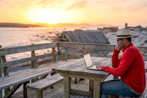 nomade digitale seduto all'aperto sulla spiaggia con un laptop da solo facendo telelavoro al tramonto - beach sunset sand wood foto e immagini stock