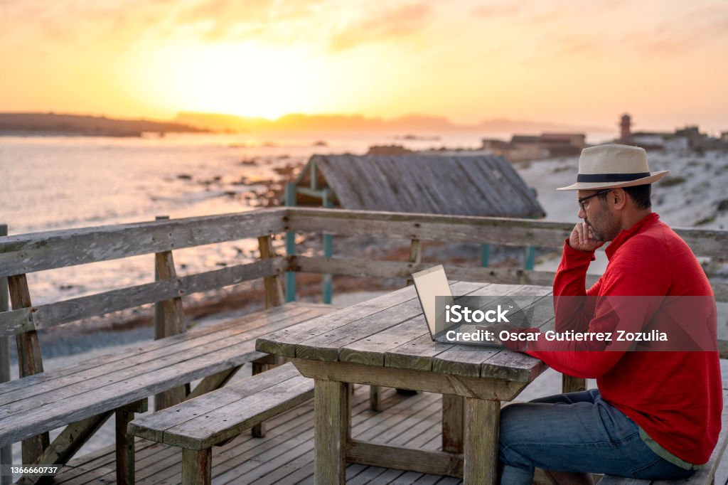 digital nomad sitting outdoors on the beach with a laptop alone doing telecommuting at sunset Telecommuting Stock Photo
