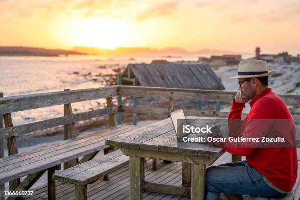 Digital Nomad Sitting Outdoors On The Beach With A Laptop Alone Doing Telecommuting At Sunset Stock Photo - Download Image Now