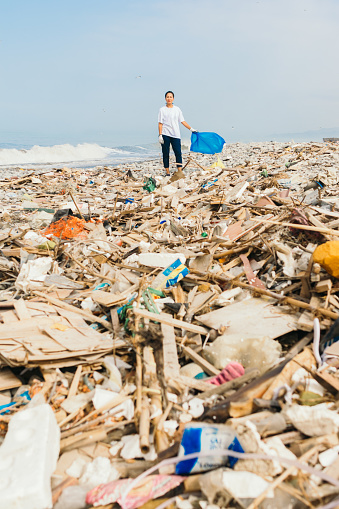 Vertical image of a latin woman holding a blue bag over a pile of garbage and trash on a polluted beach. Concept of environmental pollution