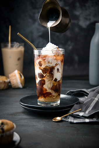 A glass filled up with cubes of ice on black background and hand pouring dark coffee on it