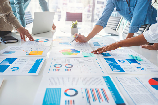 Paperwork and hands on a board room table at a business presentation or seminar. The documents have financial or marketing figures, graphs and charts on them. People are pointing to different documents. There is a laptop on the table