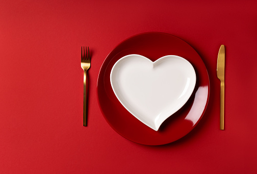 Festive table setting for valentine's day or wedding anniversary red plate and white heart shaped plate with golden cutlery on red background. Place for an inscription as a invitation card or menu.