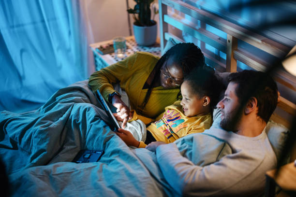 многонациональная семья, использующая цифровой планшет вместе в постели ночью. - ipad iphone smart phone ipad 3 стоковые фото и изображения