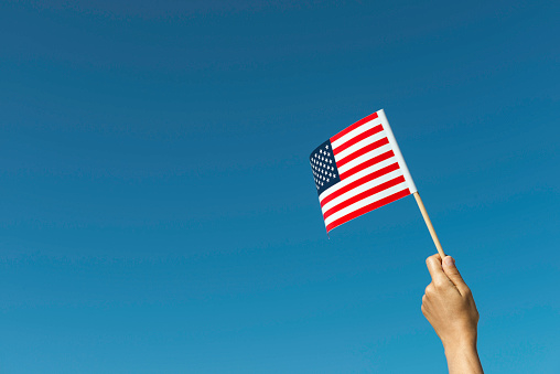 Caucasian hand who is waving American flag with clear blue sky in background.