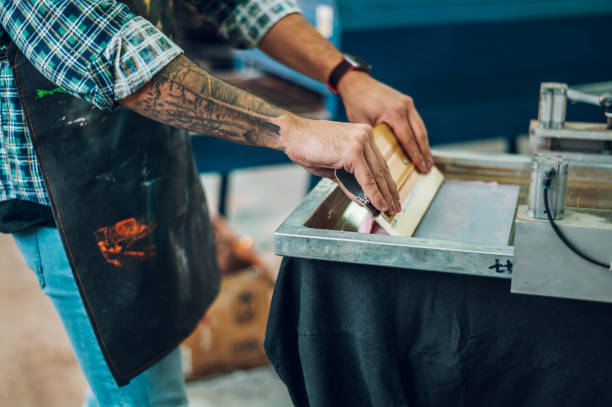 männlicher arbeiter, der tinte auf den rahmen drückt, während er die druckmaschine in einer werkstatt benutzt - silk screen fotos stock-fotos und bilder