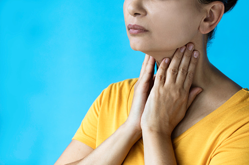 Unrecognizable female wearing a yellow t-shirt is holding her throat in pain in front of blue background.