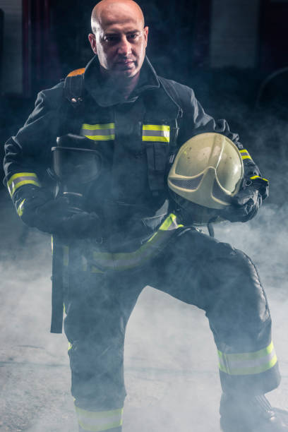 bombeiro de pé confiante segurando capacete e usando bombeiros. retrato de um bombeiro com fundo escuro com fumaça e luz azul. - turnouts - fotografias e filmes do acervo