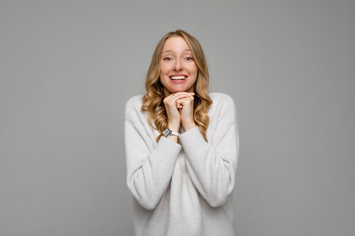 Touched, enthusiastic cute blonde woman gasping surprised and flattered, holding hands under chin, gazing camera thankful, stand pleased, wears white sweater, gray background