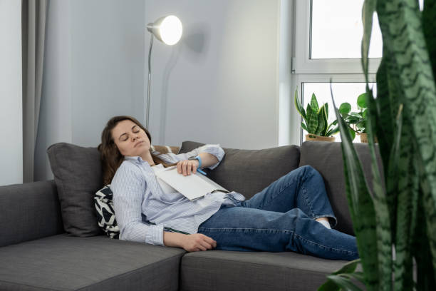 Tired woman napping on sofa at home with book on her chest, taking a break. Fatigue and sleep disorder concept stock photo