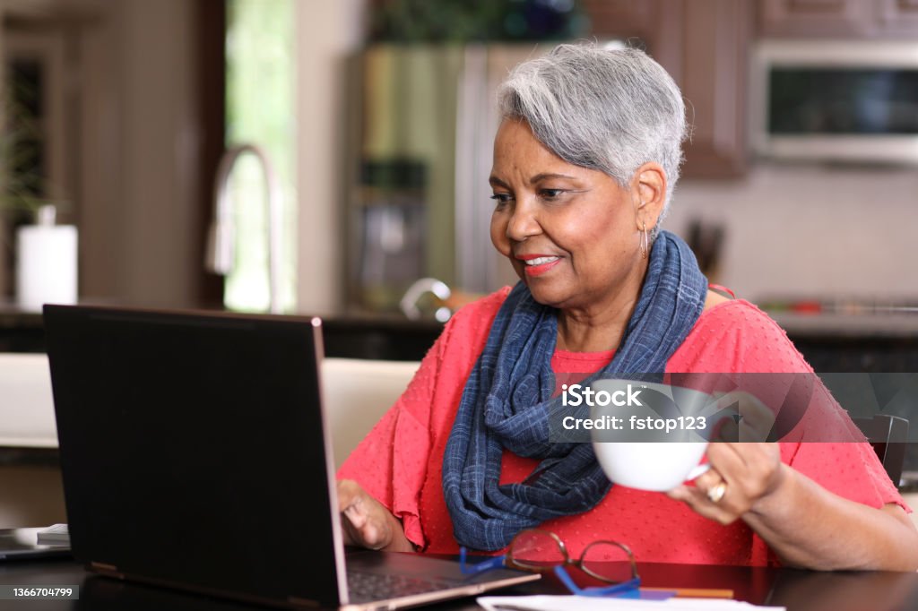 Senior adult woman using laptop computer at home. African descent, senior adult woman at home sitting at kitchen table using laptop computer. Paperwork and coffee cup also on table.  Working from home. Working At Home Stock Photo