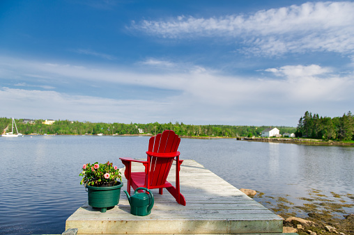 A red Adirondack chair sitting on a wooden dock in Nova Scotia, Canada. A flower pot is sitting near the chair. A boat and a building are visible in background.