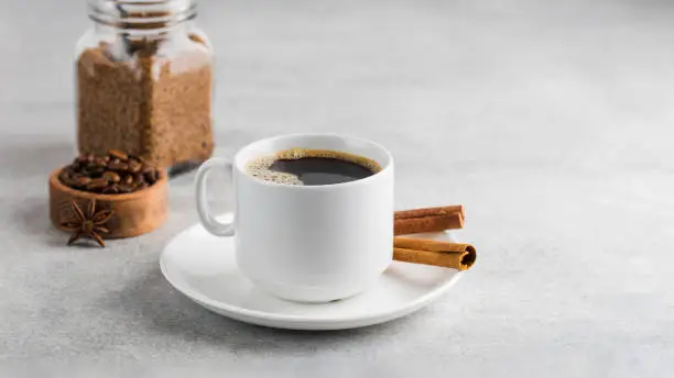 Photo of Black coffee in a white mug with cinnamon sticks on a saucer on a grey table. Copy space.