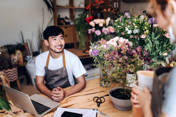 young asian couple, owners of small business flower shop, using laptop and working together in the shop against flowers and plants. discussing business matters. start-up business, business partnership and teamwork. working together for successful business - flower computer young women selling imagens e fotografias de stock