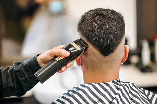 Professional barber using an electric shaver or trimmer to neaten the modern V-shaped hairstyle of a male customer viewed from the rear in close up in a barbershop
