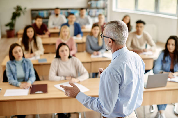Rear view of mature teacher talking to his student during lecture at university classroom. Back view of mature professor giving lecture to large group of college students in the classroom. university stock pictures, royalty-free photos & images
