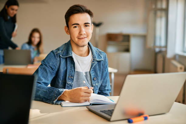 Smiling teenager taking notes while using laptop during a class at high school. Smiling high school student e-learning on laptop and writing notes in the classroom while looking at camera. teenage boys stock pictures, royalty-free photos & images