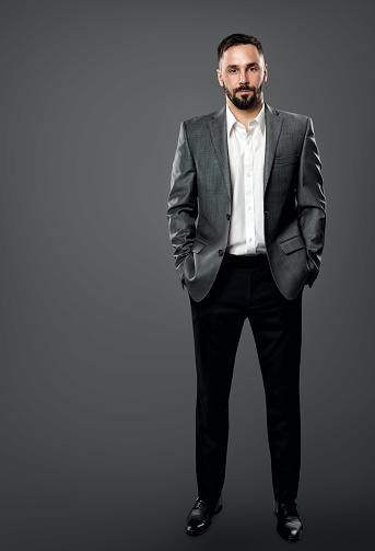 Fashion Handsome Bearded Man in Gray Suit and White Shirt without Tie. Elegant Businessman in Open Collar Shirt, Hands in Pocket Full Length over Dark Gray Studio Background