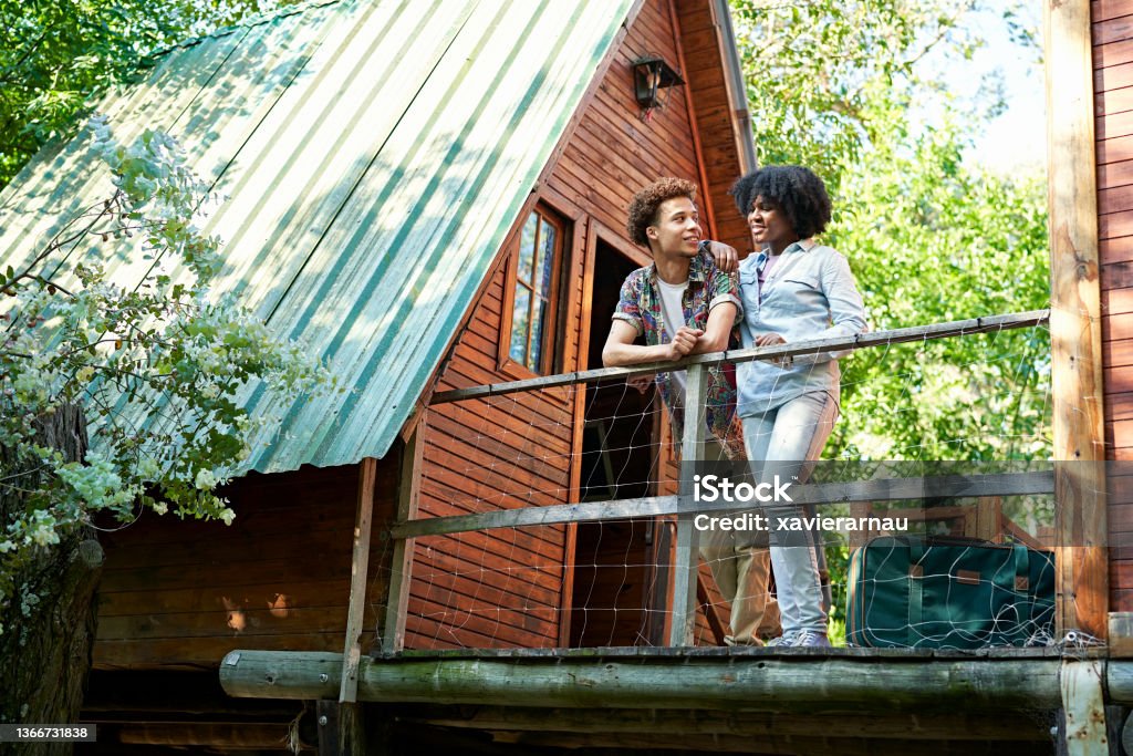 Couple standing outside cabin at start of weekend getaway Full length view of African-American man and Black woman in their 20s wearing casual clothing and enjoying view from balcony of rustic A-frame cabin. Vacation Rental Stock Photo