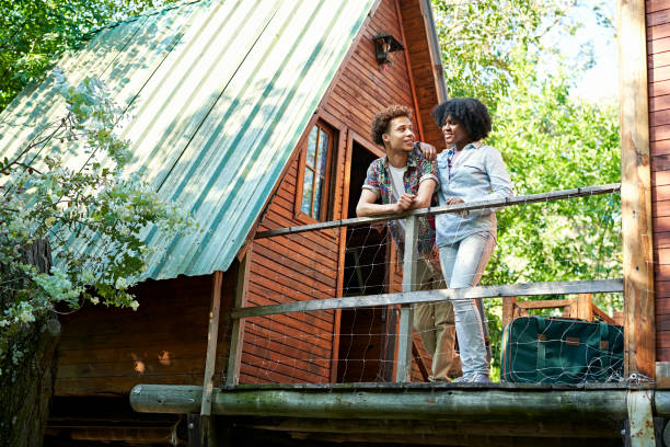 Couple standing outside cabin at start of weekend getaway Full length view of African-American man and Black woman in their 20s wearing casual clothing and enjoying view from balcony of rustic A-frame cabin. holiday villa stock pictures, royalty-free photos & images