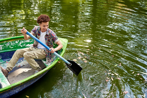 Elevated full length view of man in casual clothing holding paddle and laughing as he tries to steer boat on water.