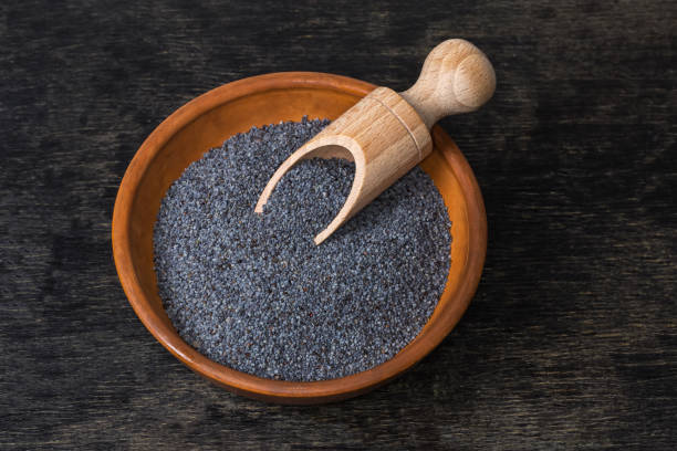 Dry raw poppy seeds of blue variety in the clay bowl with wooden spice spoon on a black surface