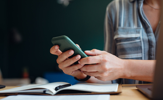 Anonymous woman sitting at her desk at home. She has her notebook and laptop in front of her. She is holding her phone and texting.