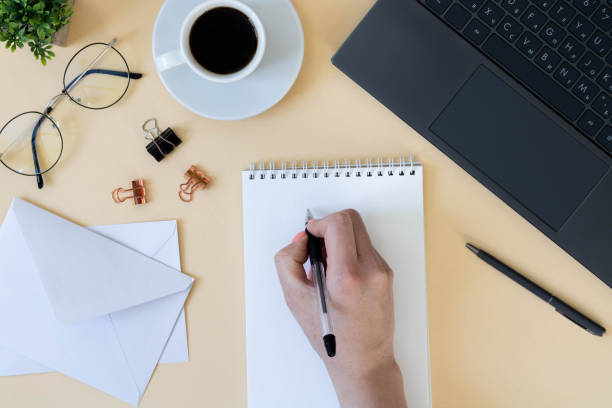 istock Women's hands write in a notebook. Top view of office desk with laptop, notebook and coffee cup, envelopes, stationery. Workplace, flat lay, copy space. 1366743944