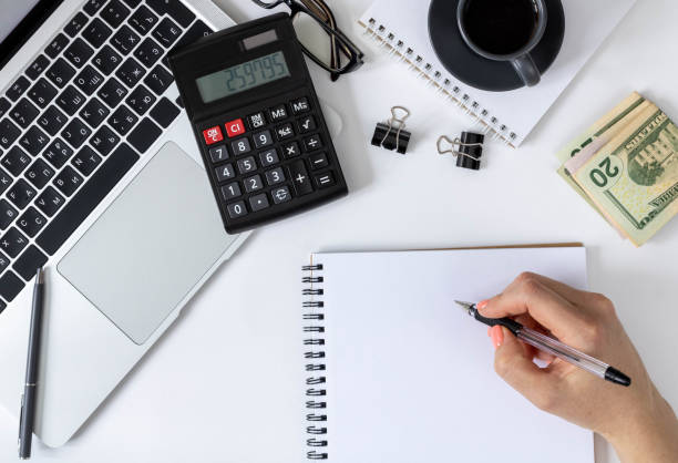 istock Female hands write in a spiral planner. Workspace: laptop, calculator, money and coffee on a white background, top view. 1366743948