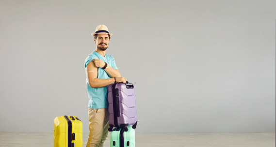 Vacation, new travel rules and vaccination against coronavirus covid-19. Caucasian millennial man traveler standing with luggage suitcase show arm with patch. Studio portrait on grey copy space