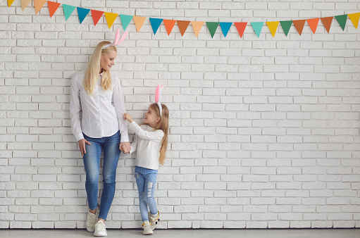 Happy easter. Mother and daughter with rabbit ears are standing smiling against a white brick wall.