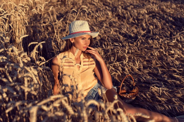A blonde woman in a blue plaid shirt and jeans stands among barley ears in a field, stroking the ears. She looks towards the sunset across the field.