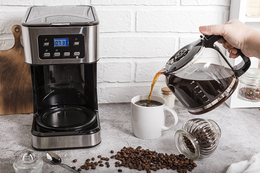 A woman's hand pours coffee from a coffee pot into a cup. Home life. Electric coffee maker on the kitchen countertop