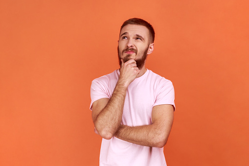 Portrait of thoughtful handsome bearded man holding his chin and pondering idea, confused not sure about solution, wearing pink T-shirt. Indoor studio shot isolated on orange background.